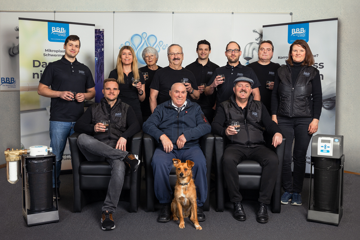 Das Team von BBB posiert in Firmenkleidung mit Wasser in der Hand. Im Vordergrund sitzt ein Hund. Links und rechts im Bild stehen Wasserfiltersysteme.